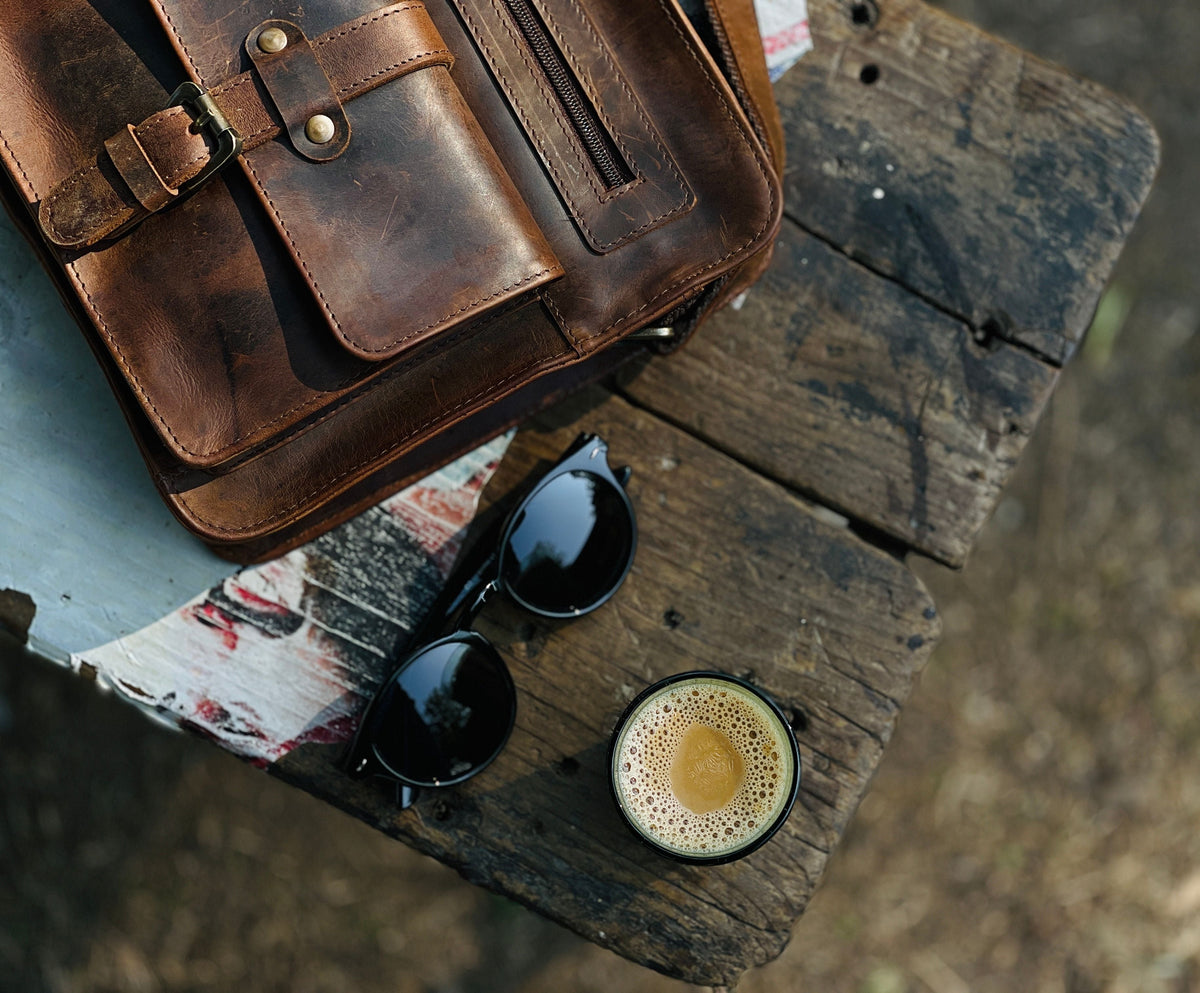 Détail d’un sac en cuir brun aux nuances marquées, posé sur une table en bois à côté de lunettes et d’un café.
